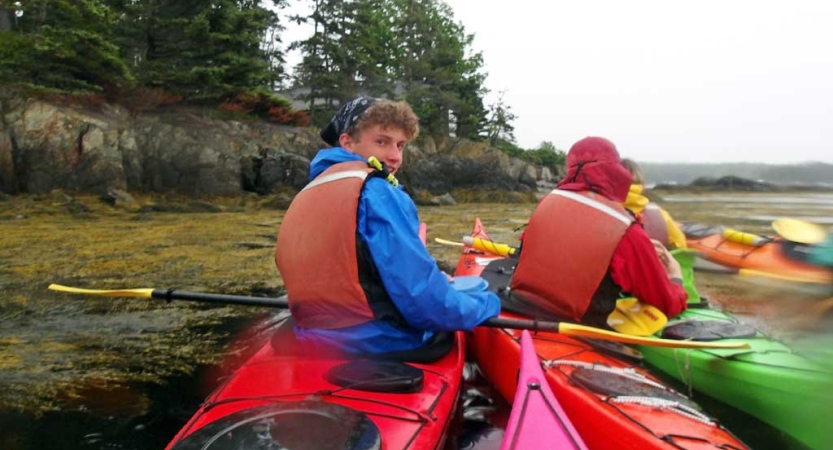 Students sit in colorful kayaks near a rocky shore. 